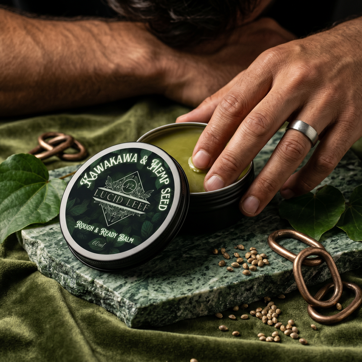 A tin f Lucid Leef Kawakawa and Hemp Seed Rough and Ready Balm sitting on a green stone slab with the lid removed to the side. The stone slab sits on a green crushed velvet cloth adorned with kawakawa leaves, hemp seeds and bronze chain links. The hand of a male model is scooping some balm from the tin with his fingertip.