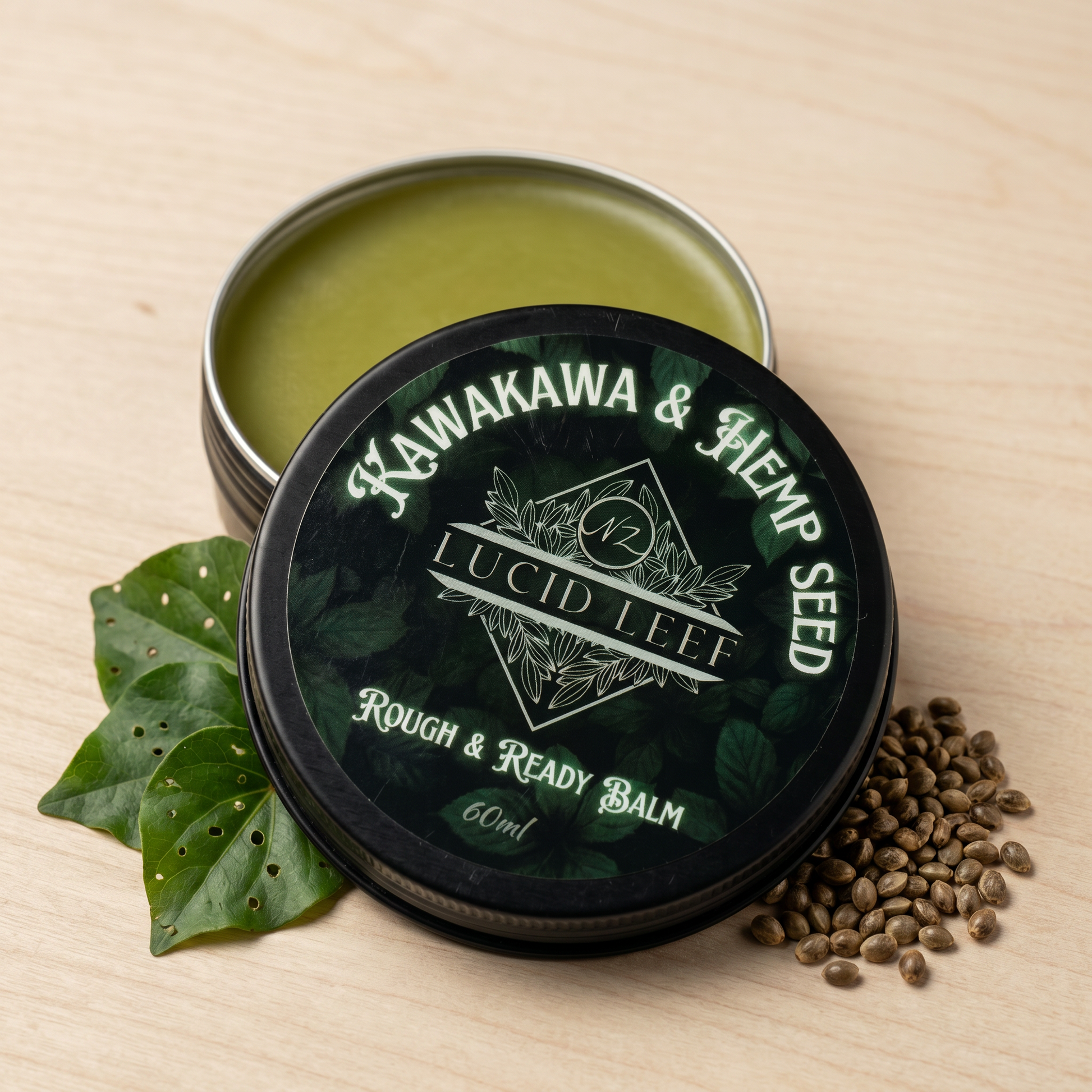 A tin of Lucid Leef Kawakawa and Hemp Seed Rough and Ready Balm sitting flat on a pale woodgrain surface with the lid partially removed to show the balm inside. To the left of the tin are 3 Kawakawa leaves with small bug holes and to the right, a small group of hemp seeds.