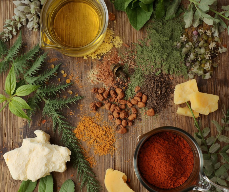 Spices, herbs, and a jar of oil on a wooden surface with greenery.